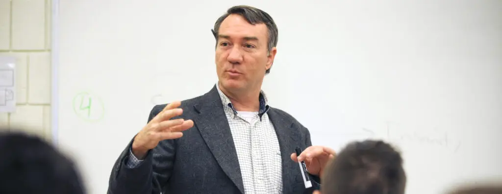 A man in a blazer gestures with his hands while speaking in front of a whiteboard during a classroom lecture. Several students are partially visible in the foreground.