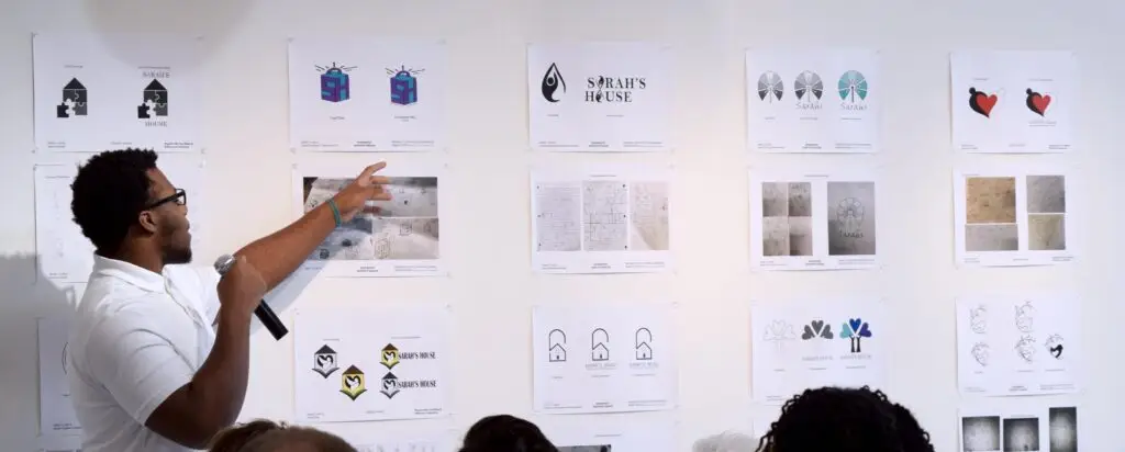 A man holding a microphone points to design sketches and drawings displayed on a white wall during a presentation, with several people seated and watching.