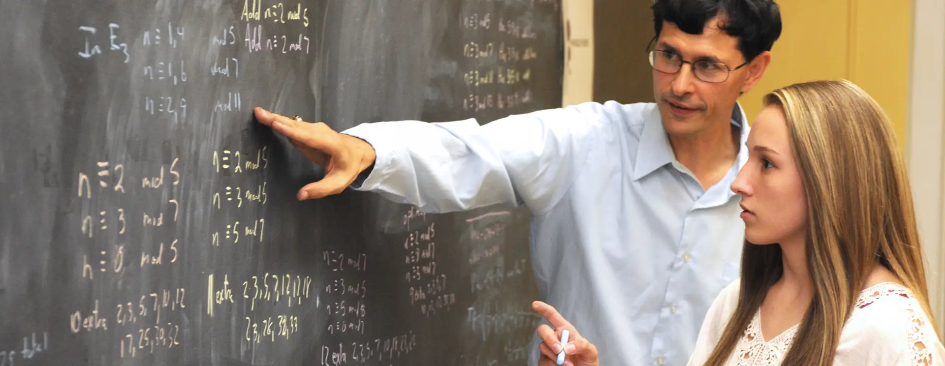 A teacher explains math equations on a blackboard to a student, who is holding a piece of chalk and listening attentively. Both are focused on the board, which is filled with handwritten formulas.