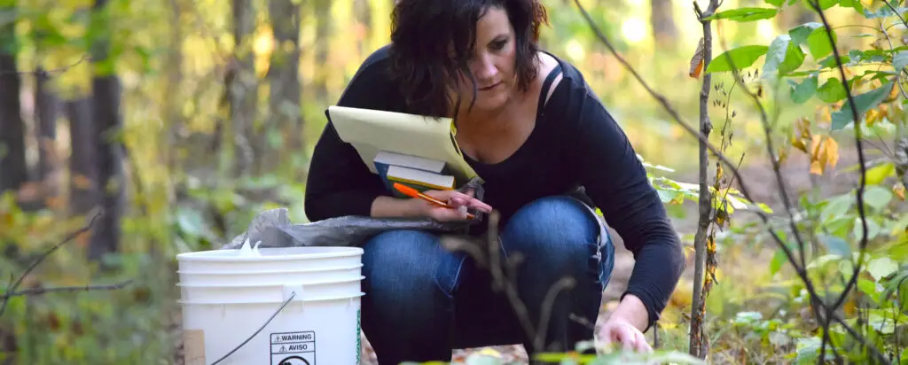 A woman crouches in a forest, holding notebooks and a pencil, and reaching toward the ground near a white bucket. Sunlight filters through the trees and green foliage surrounds her.