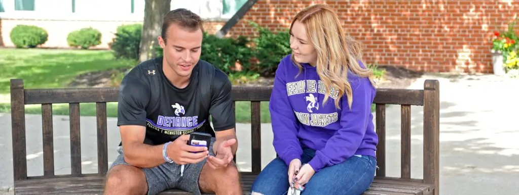 Two college students sit on a wooden bench outdoors. The man shows something on his phone to the woman, who is looking at him. Both wear Defiance College shirts, and there are trees and a brick building in the background.