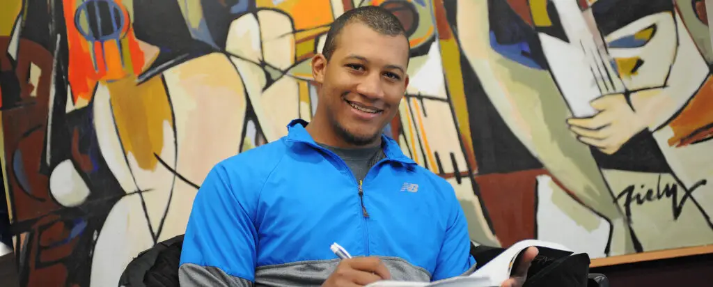 A young man in a blue jacket smiles while writing in a notebook. He is seated in front of a colorful abstract mural.