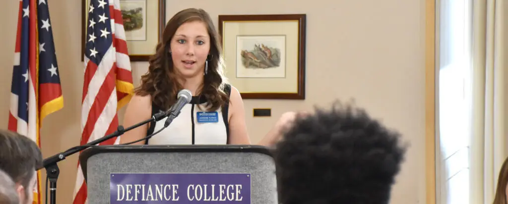 A woman stands at a podium labeled Defiance College, speaking into a microphone. She faces an audience, with framed pictures and American flags visible in the background.