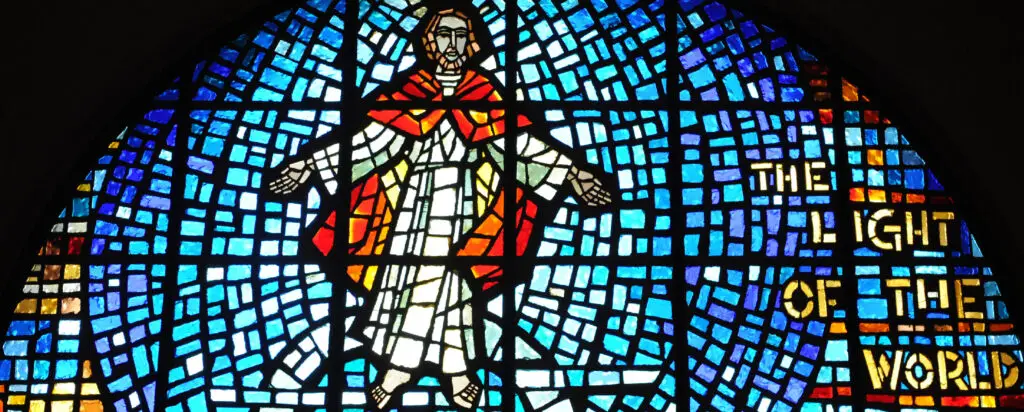 Stained glass window depicting a figure in white and red robes with arms outstretched, surrounded by blue and yellow glass tiles. The words “The Light of the World” appear on the right side.
