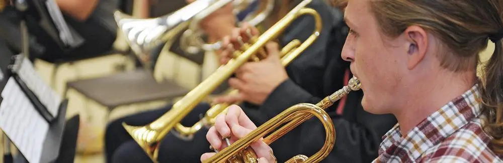 Several people play brass instruments, such as trumpets, in what appears to be a band or orchestra rehearsal. The focus is on a person in the foreground wearing a plaid shirt, with sheet music visible to the side.