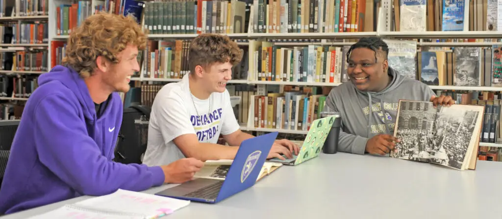 Three college students sit at a library table with laptops and notebooks, smiling and talking; one student holds up an open book with a black-and-white historical image. Bookshelves are visible in the background.