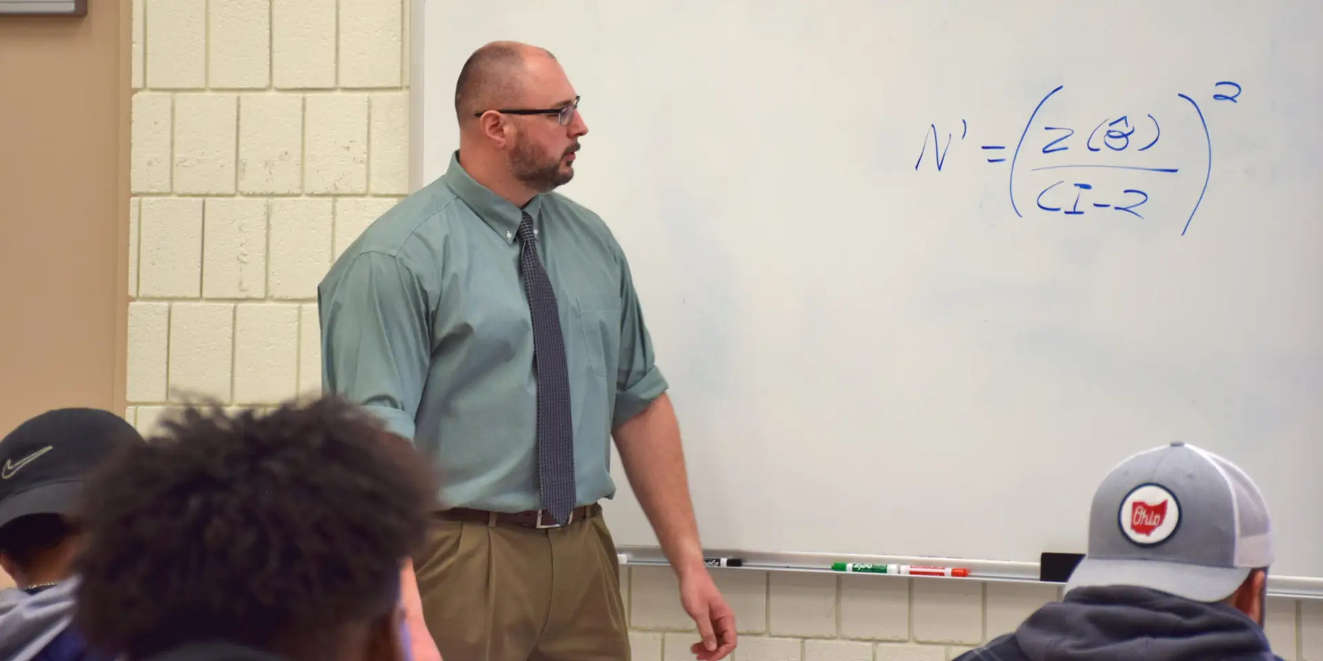 A man in a green shirt and tie stands next to a whiteboard with a mathematical formula written on it, teaching a class with students visible in the foreground.