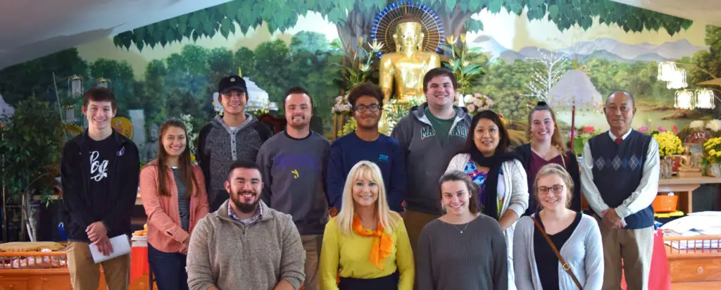 A group of fifteen people pose and smile indoors in front of a golden Buddha statue, with a painted green landscape mural behind them. The group is casually dressed and standing in two rows.
