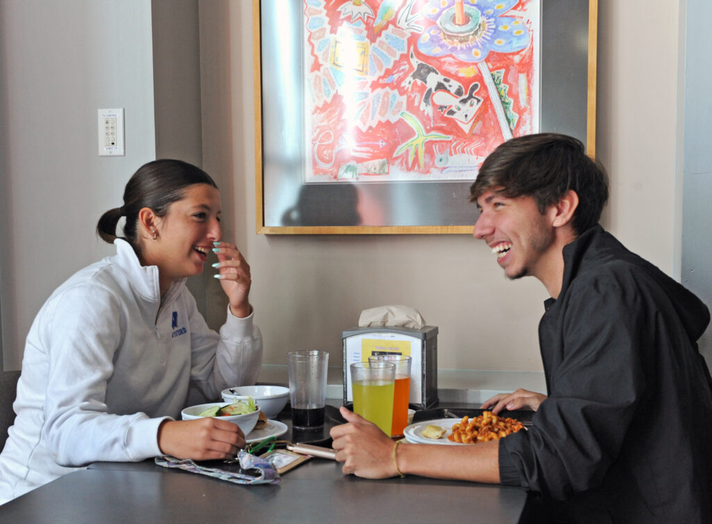 Two young people sit at a table, smiling and laughing while enjoying a meal together. There are drinks and plates of food in front of them, and colorful artwork hangs on the wall behind them.