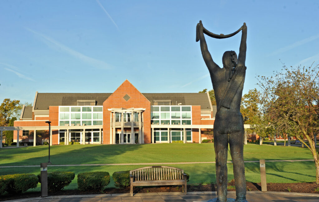 A bronze statue of a person holding a cloth overhead stands in front of a modern brick and glass building with a green lawn and trimmed bushes, under a clear blue sky.