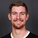 A young man with short light brown hair, a trimmed beard, and a friendly smile poses in front of a plain dark background. He is wearing a black shirt with a white collar.