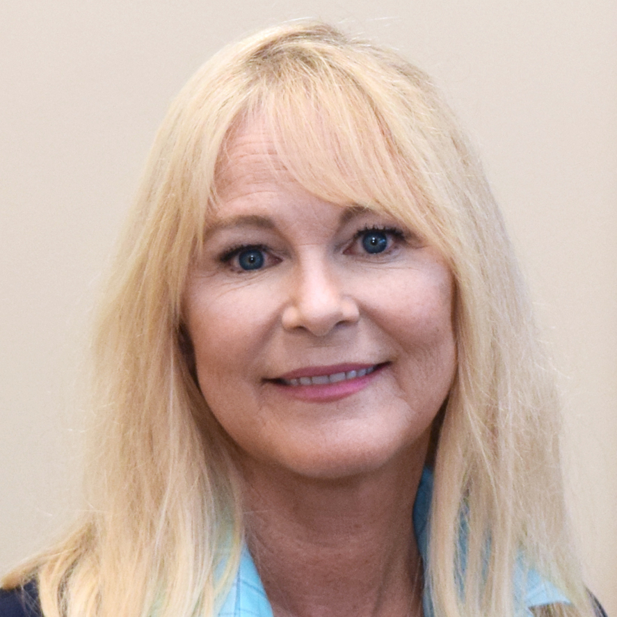 A woman with long blonde hair and blue eyes smiles softly at the camera. She is wearing a light blue collared shirt and a dark jacket, standing against a plain, light-colored background.