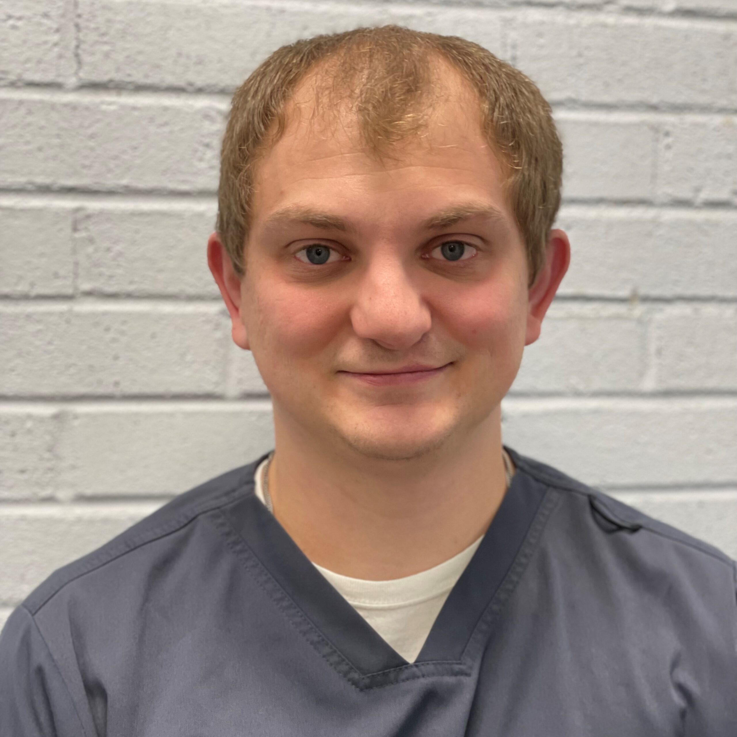 A young man with short light brown hair wearing a gray scrub top and white undershirt is smiling slightly, standing in front of a white brick wall.