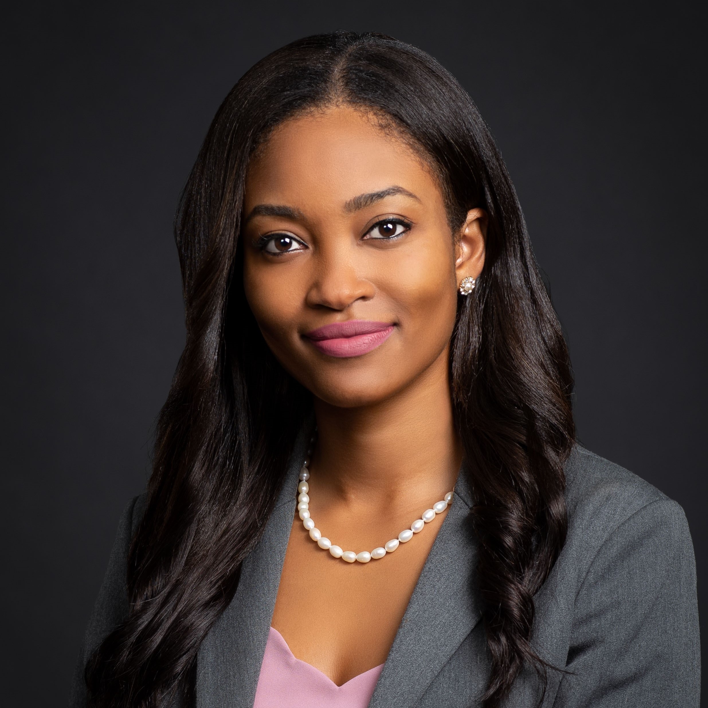 A woman with long, dark hair wearing a gray blazer, pink top, pearl necklace, and stud earrings smiles slightly against a plain dark background.
