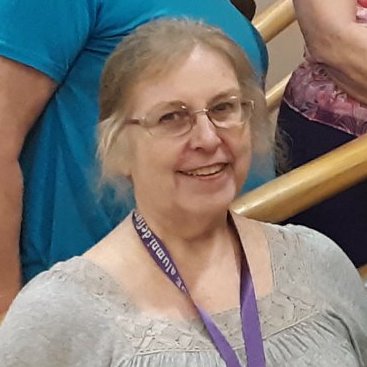An older woman with glasses and gray hair pulled back smiles at the camera. She is wearing a light gray top and a purple lanyard around her neck. Other people are partially visible in the background.