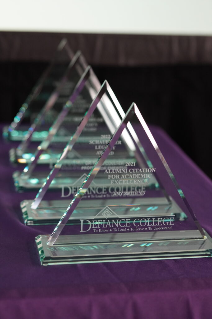Five glass triangular awards are displayed in a row on a purple tablecloth. The front award reads “Defiance College” with inscriptions for “Alumni Citation for Academic Excellence.”.
