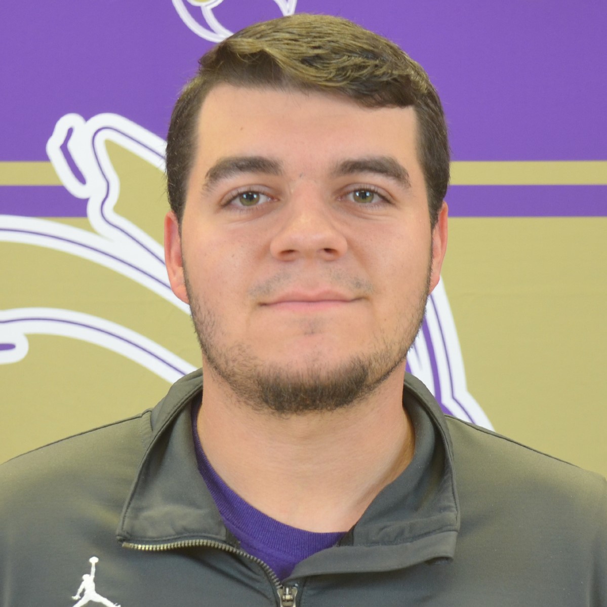 A young man with short dark hair and a trimmed beard is wearing a gray jacket. He is posing in front of a purple and gold background with a stylized graphic design.