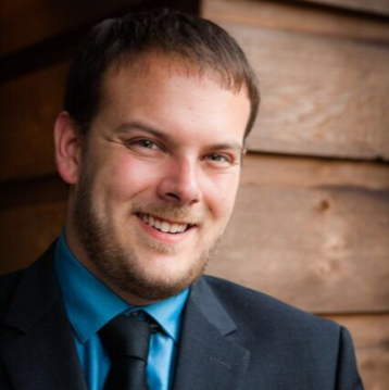 A man with short brown hair and a beard, wearing a dark suit, blue shirt, and black tie, smiles while standing in front of a wooden wall.