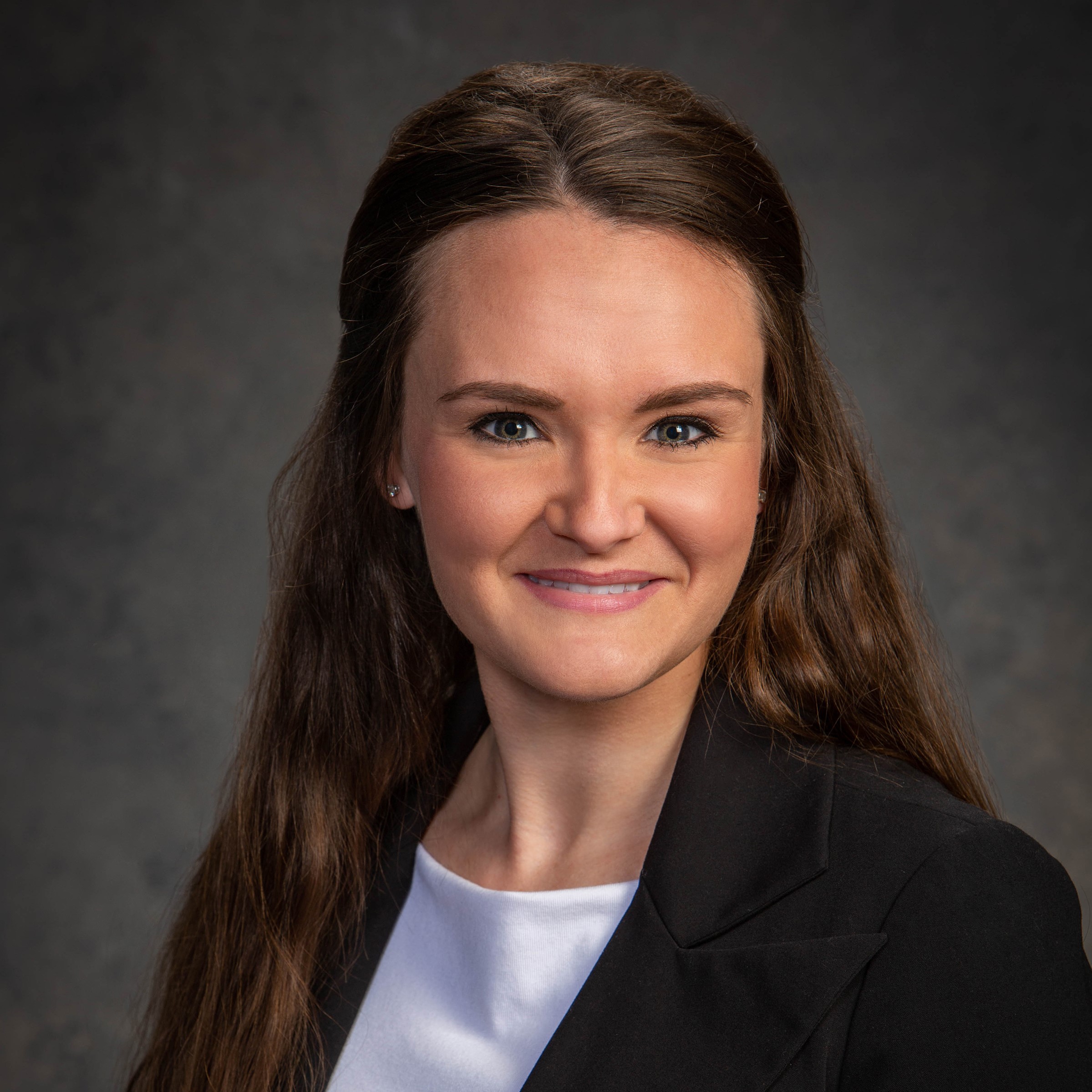 Professional headshot of a woman with long brown hair, wearing a black blazer and white top, smiling in front of a dark, neutral background.