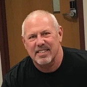 A middle-aged man with short white hair and a trimmed beard, wearing a black shirt, smiling indoors near a wooden door.