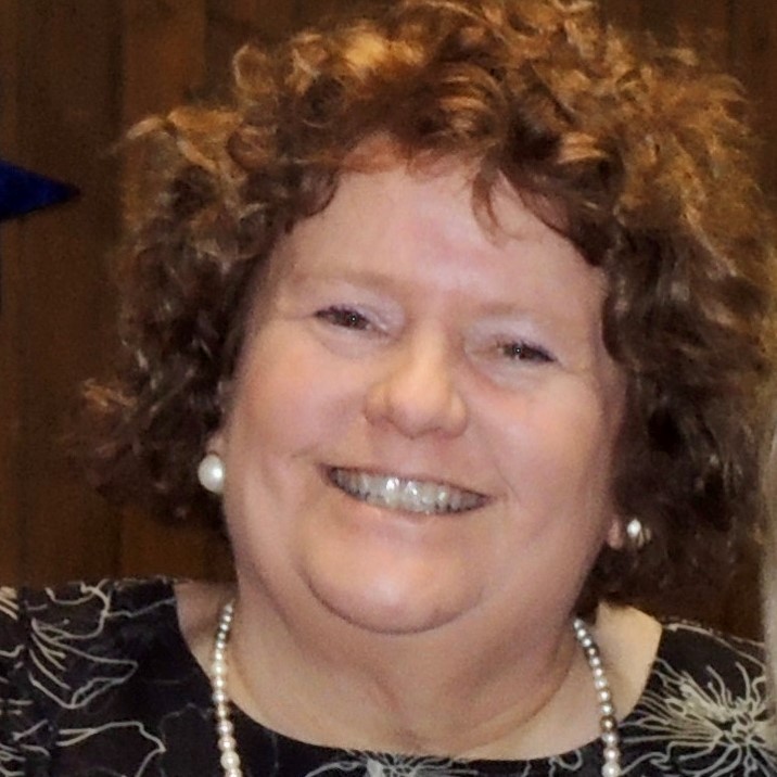 A woman with curly brown hair and light skin smiles at the camera. She is wearing a dark top with a light pattern, pearl earrings, and a pearl necklace. The background is out of focus wood paneling.