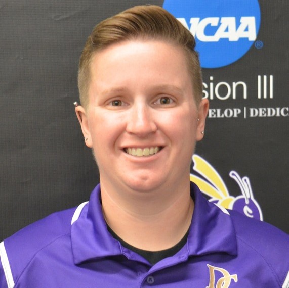 A person with short, light brown hair, wearing a purple collared shirt with a yellow and white logo, smiles in front of a black backdrop displaying the NCAA Division III logo.