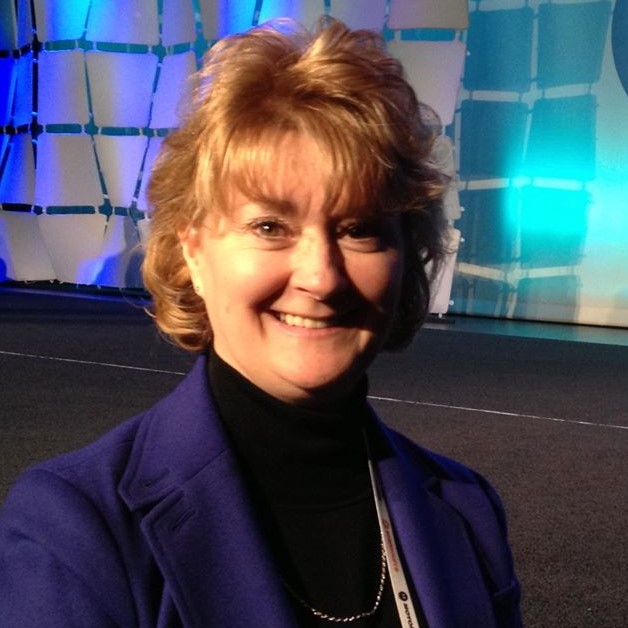 A woman with short, curly blonde hair wearing a blue blazer and black turtleneck smiles at the camera. She stands in front of a modern, blue-lit stage backdrop.
