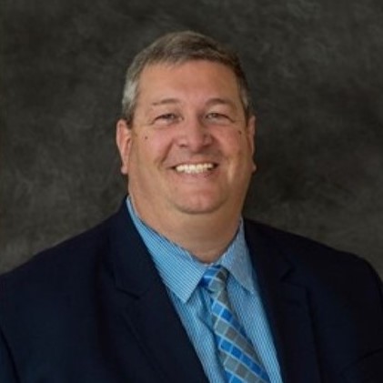 A middle-aged man with short gray hair smiles while wearing a dark suit jacket, light blue striped shirt, and blue patterned tie against a dark gray background.