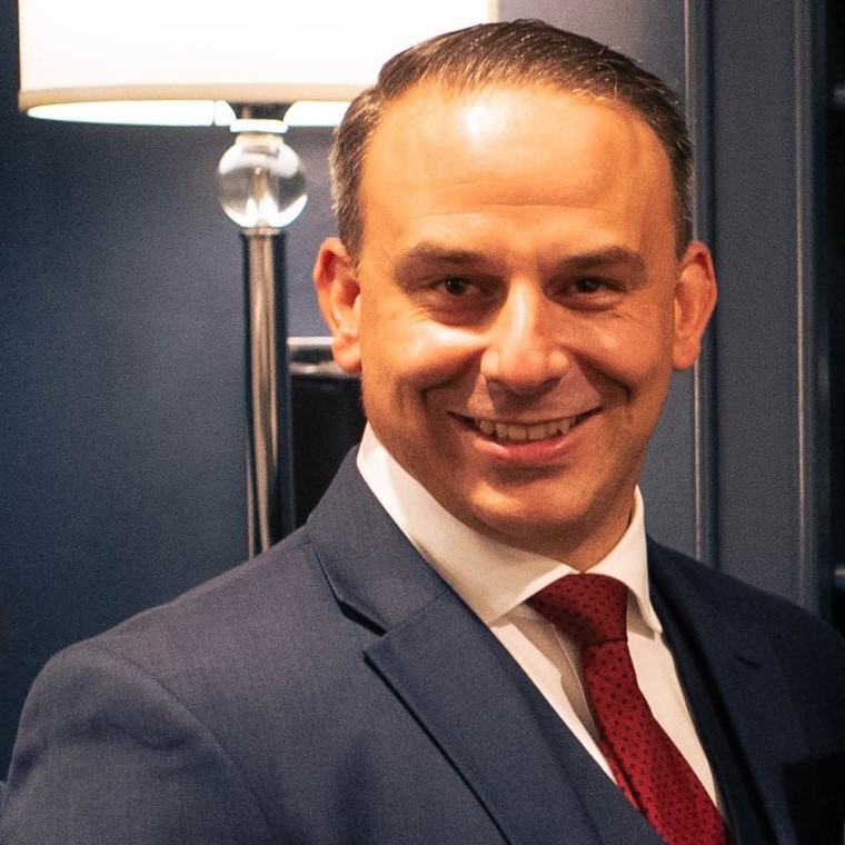 A man in a navy blue suit, white shirt, and red tie smiles at the camera. He has short dark hair and stands in front of a lamp and a dark-colored wall.