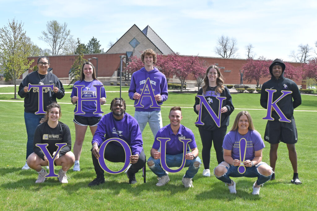 Nine smiling students pose outdoors on grass, wearing purple shirts. They each hold large letters that together spell “THANK YOU!” Trees and a building are visible in the background.