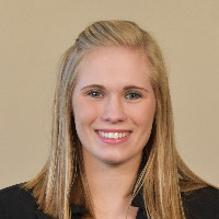 A young woman with straight blonde hair, wearing a black blazer and smiling, is posed in front of a plain beige background.