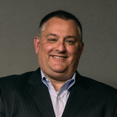 A smiling man wearing a dark pinstripe suit jacket and a light blue collared shirt, posed against a plain gray background.