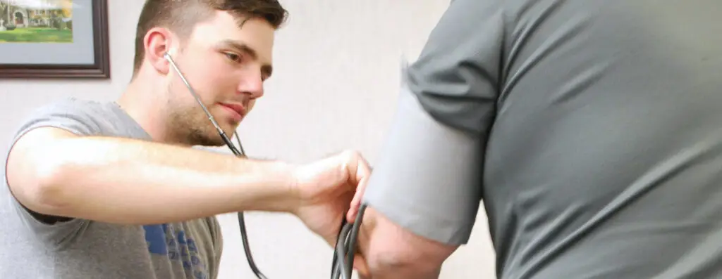 A healthcare worker uses a stethoscope and blood pressure cuff to measure a patient’s blood pressure. The patient is seated with their arm extended, and the worker is focused on the task.