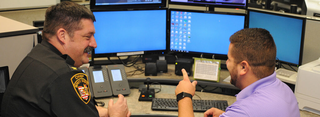 A police officer and another man sit at a desk with multiple computer monitors, having a friendly conversation and smiling in an office setting.