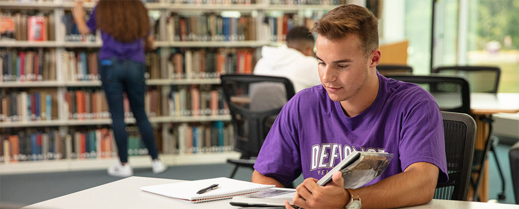 A student wearing a purple shirt sits at a desk in a library, holding a book and writing in a notebook. Bookshelves filled with books and another person browsing the shelves are visible in the background.