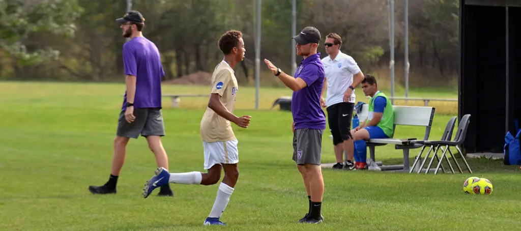 A soccer player in a gold jersey talks to a coach in purple on a grassy field. Other team members and staff sit or stand near a bench, with a soccer ball and sports equipment nearby.