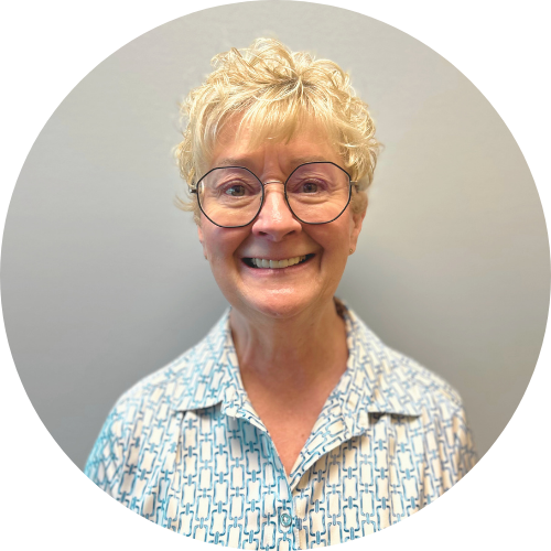 Smiling older woman with short curly blonde hair and glasses, wearing a patterned button-up shirt, standing in front of a plain light background.