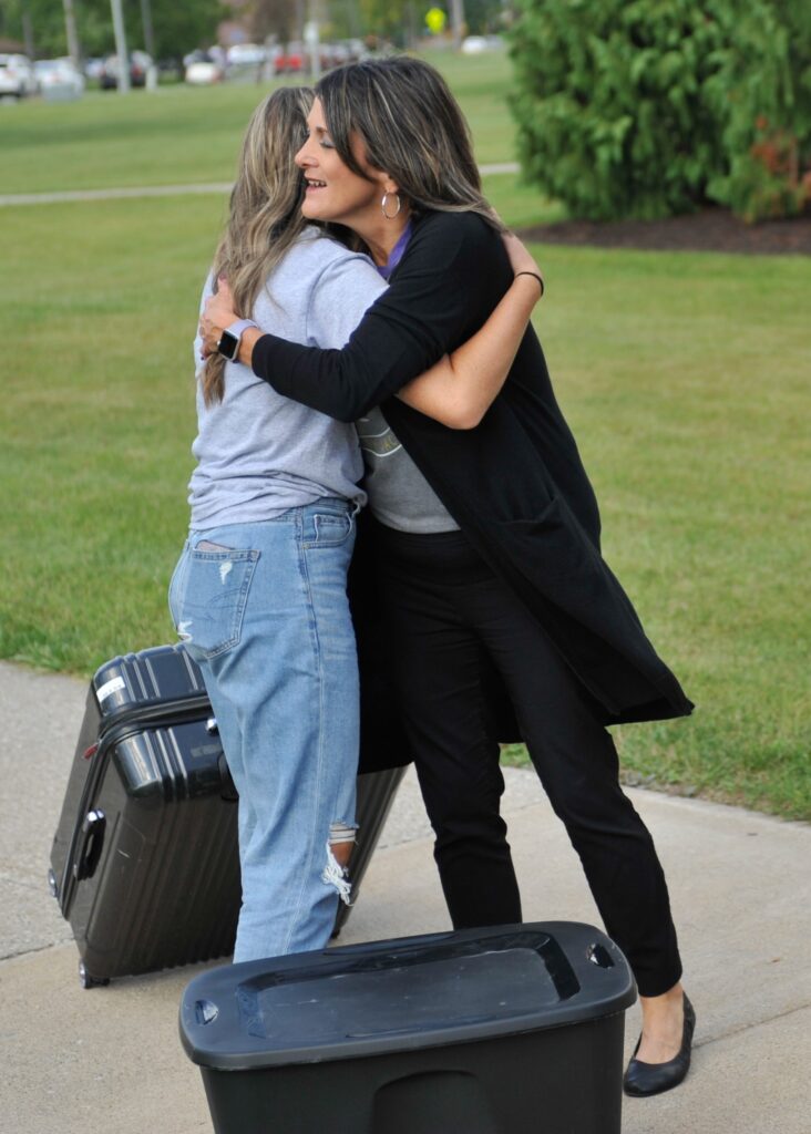 Two women hug on a sidewalk near grass, one holding a suitcase and wearing jeans, the other in black pants and a cardigan. A large plastic storage bin sits in the foreground.