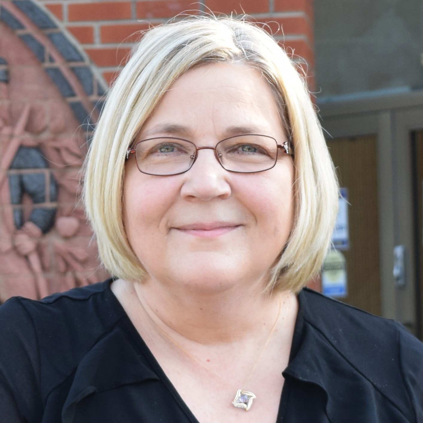 A woman with short blonde hair and glasses, wearing a black top and a square pendant necklace, stands outdoors in front of a brick wall and glass door.