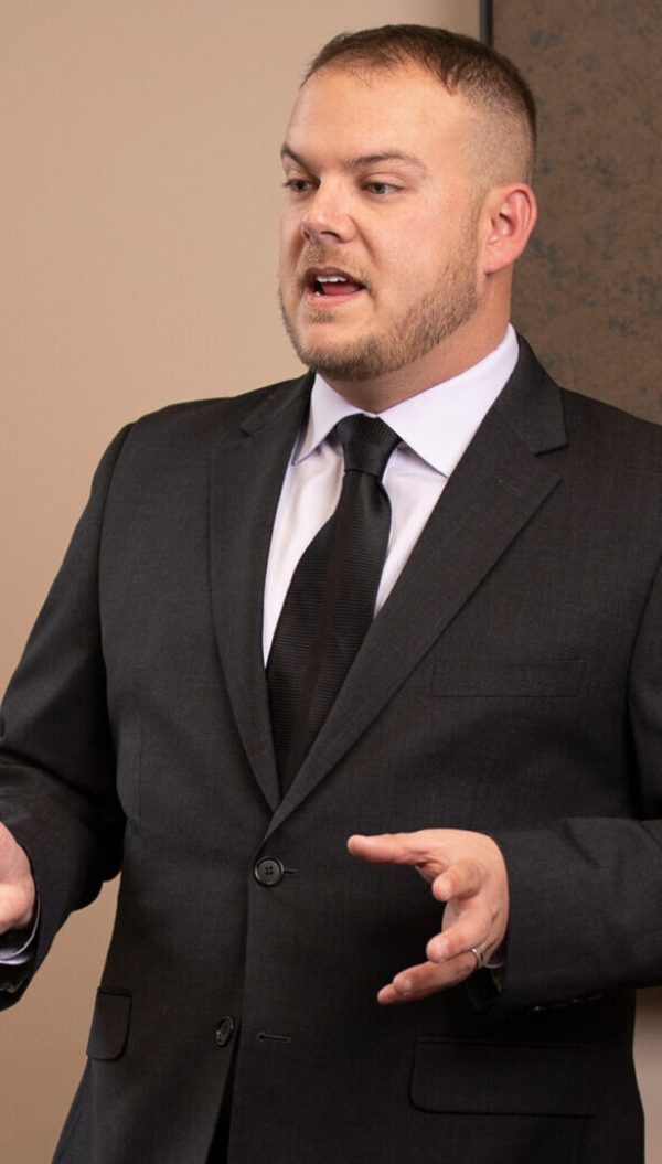 A man in a dark suit and black tie speaks while gesturing with his hands. He stands in front of a neutral-colored background, appearing engaged and confident.
