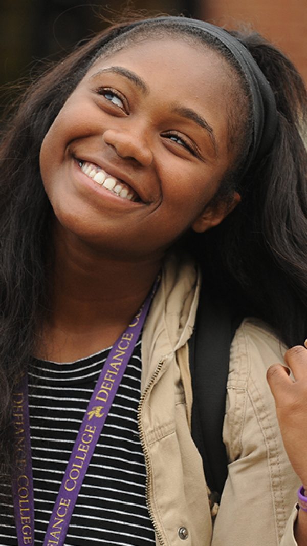 A young woman with long dark hair smiles joyfully, looking upward. She wears a striped shirt, tan jacket, and a purple lanyard around her neck, and stands outdoors with a blurred background.